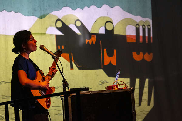 Sulla sinistra una ragazza canta al microfono e suona una chitarra elettrica illuminata da una luce tenue rossastra. Sullo sfondo campeggia su tutta la parete un'opera stilizzata che ritrae un paesaggio con un carro armato.